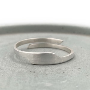 May include: A silver-colored wrap ring with a brushed metal finish. The ring has a unique, asymmetrical design, with one end wrapping around the finger. The ring is shown on a gray surface.