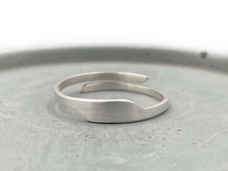 May include: A silver-colored wrap ring with a brushed metal finish. The ring has a unique, asymmetrical design, with one end wrapping around the finger. The ring is shown on a gray surface.
