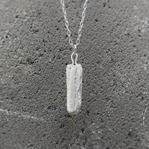 Puede incluir: Un collar con colgante plateado con un colgante rectangular texturizado. El colgante cuelga de una delicada cadena. El collar se muestra sobre un fondo gris oscuro texturizado. El colgante mide unos 2 cm.