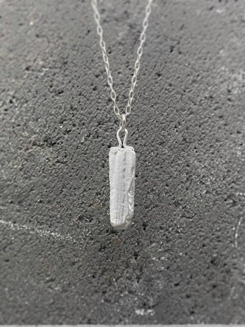 Puede incluir: Un collar con colgante plateado con un colgante rectangular texturizado. El colgante cuelga de una delicada cadena. El collar se muestra sobre un fondo gris oscuro texturizado. El colgante mide unos 2 cm.