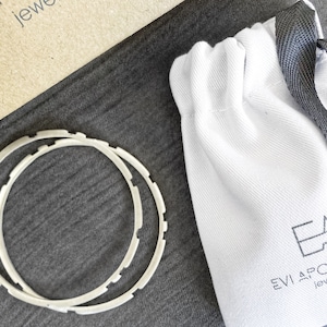May include: Two silver bangle bracelets with a textured design, packaged in a white drawstring bag with a black ribbon and the logo "Evi Apostolo Jewellery".