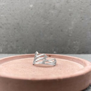 Pode incluir: Um anel de prata com um design geom&eacute;trico. O anel est&aacute; sobre uma superf&iacute;cie de cor terracota.