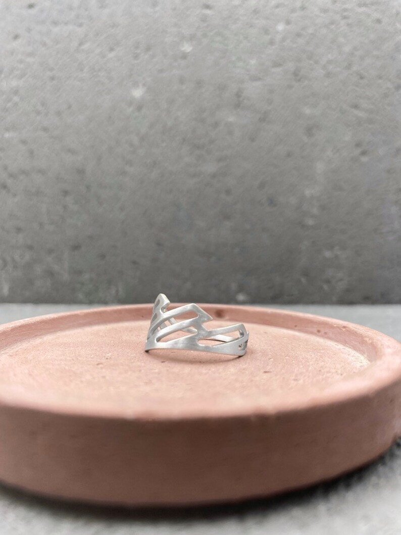 Pode incluir: Um anel de prata com um design geom&eacute;trico. O anel est&aacute; sobre uma superf&iacute;cie de cor terracota.