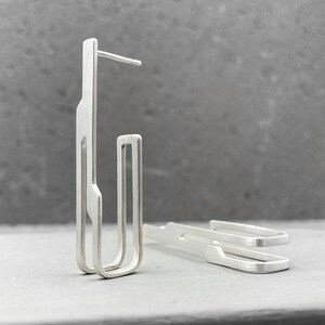 May include: A pair of silver geometric earrings with a modern design. The earrings are made of thin metal and have a simple, minimalist style.
