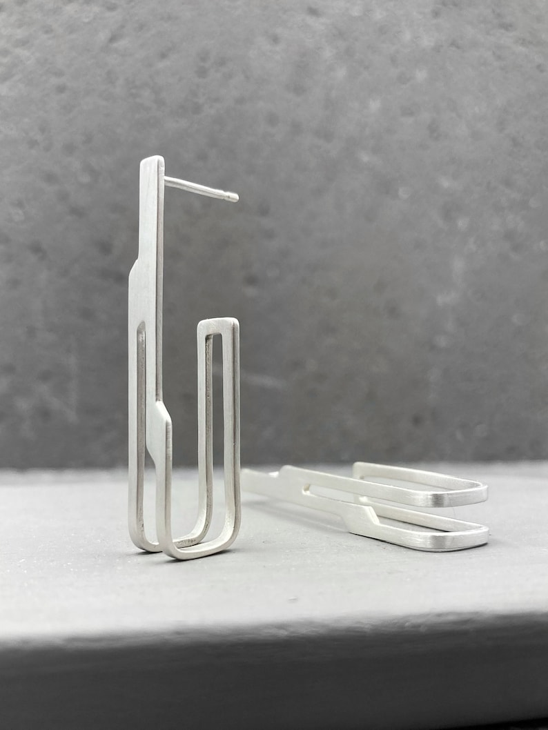 May include: A pair of silver geometric earrings with a modern design. The earrings are made of thin metal and have a simple, minimalist style.