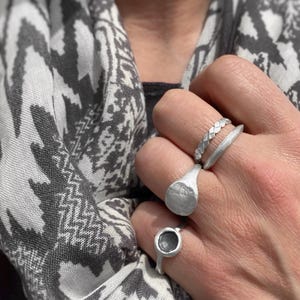 May include: A set of three silver-toned rings worn on fingers. The top ring features a braided texture, the middle ring has a smooth, thick band with a flat circular face, and the bottom ring is a thin band with a small, dark, circular inset.