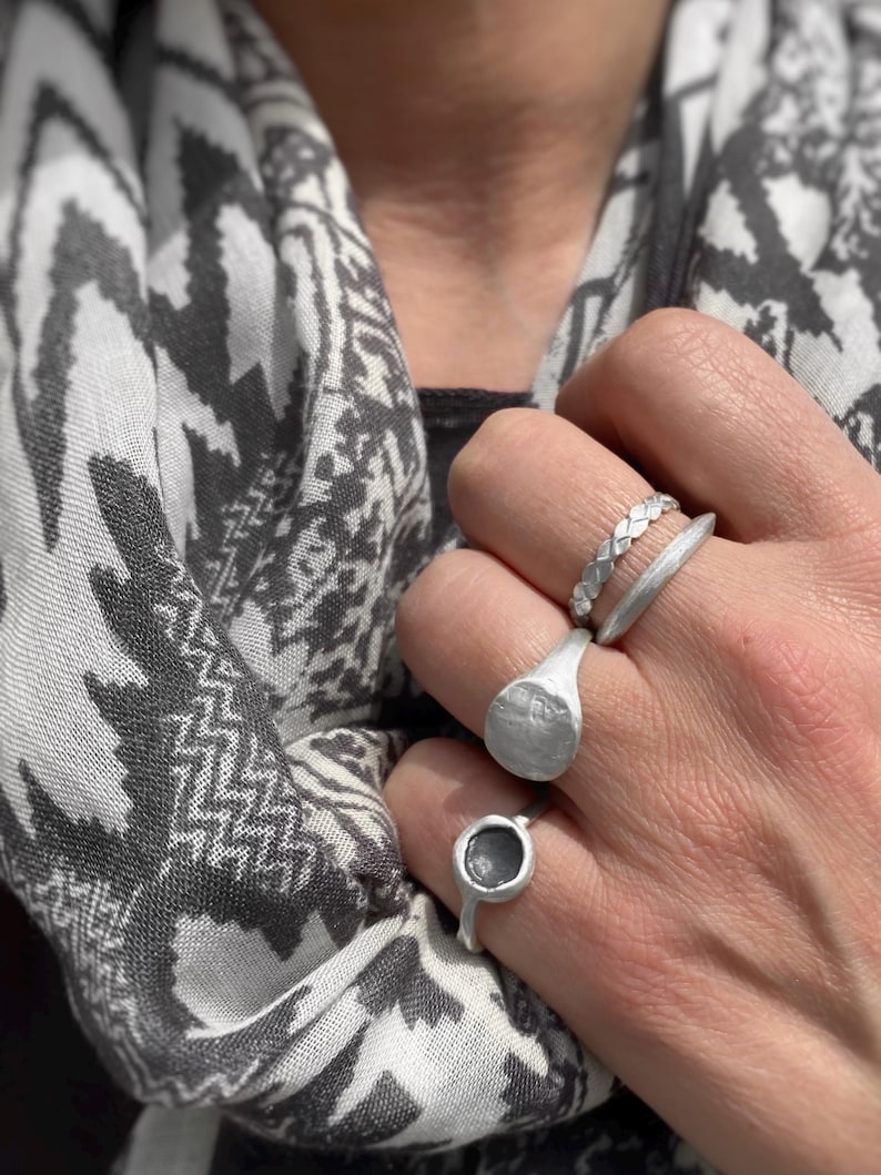 May include: A set of three silver-toned rings worn on fingers. The top ring features a braided texture, the middle ring has a smooth, thick band with a flat circular face, and the bottom ring is a thin band with a small, dark, circular inset.