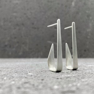 Può includere: Un paio di orecchini d'argento con un design geometrico unico. Gli orecchini sono realizzati in un unico pezzo di metallo piegato in una forma curva.