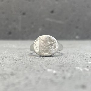 May include: A silver-toned signet ring featuring a circular face with a textured, uneven surface. The metal has a matte finish with visible indentations and markings across the round top, tapering into a smooth, solid band that encircles the finger.