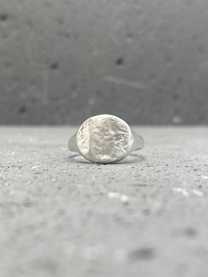 May include: A silver-toned signet ring featuring a circular face with a textured, uneven surface. The metal has a matte finish with visible indentations and markings across the round top, tapering into a smooth, solid band that encircles the finger.