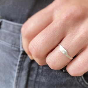 May include: A silver ring with a geometric design. The ring is worn on a finger, and the design features a rectangular shape with angled edges. The ring appears to be made of a brushed metal.