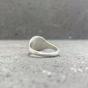 May include: A silver-toned signet ring with a smooth, matte finish. The ring features a rounded band and a flat, circular face oriented upwards. The metal surface has a consistent, non-reflective appearance throughout the entire piece of jewelry.