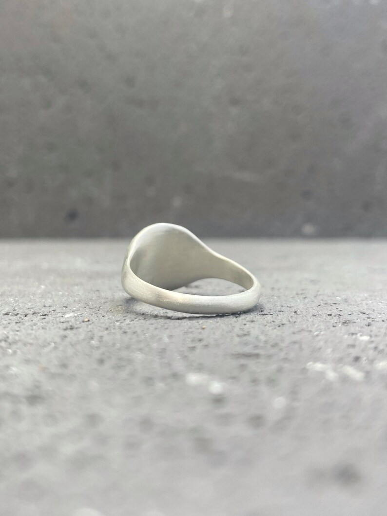 May include: A silver-toned signet ring with a smooth, matte finish. The ring features a rounded band and a flat, circular face oriented upwards. The metal surface has a consistent, non-reflective appearance throughout the entire piece of jewelry.