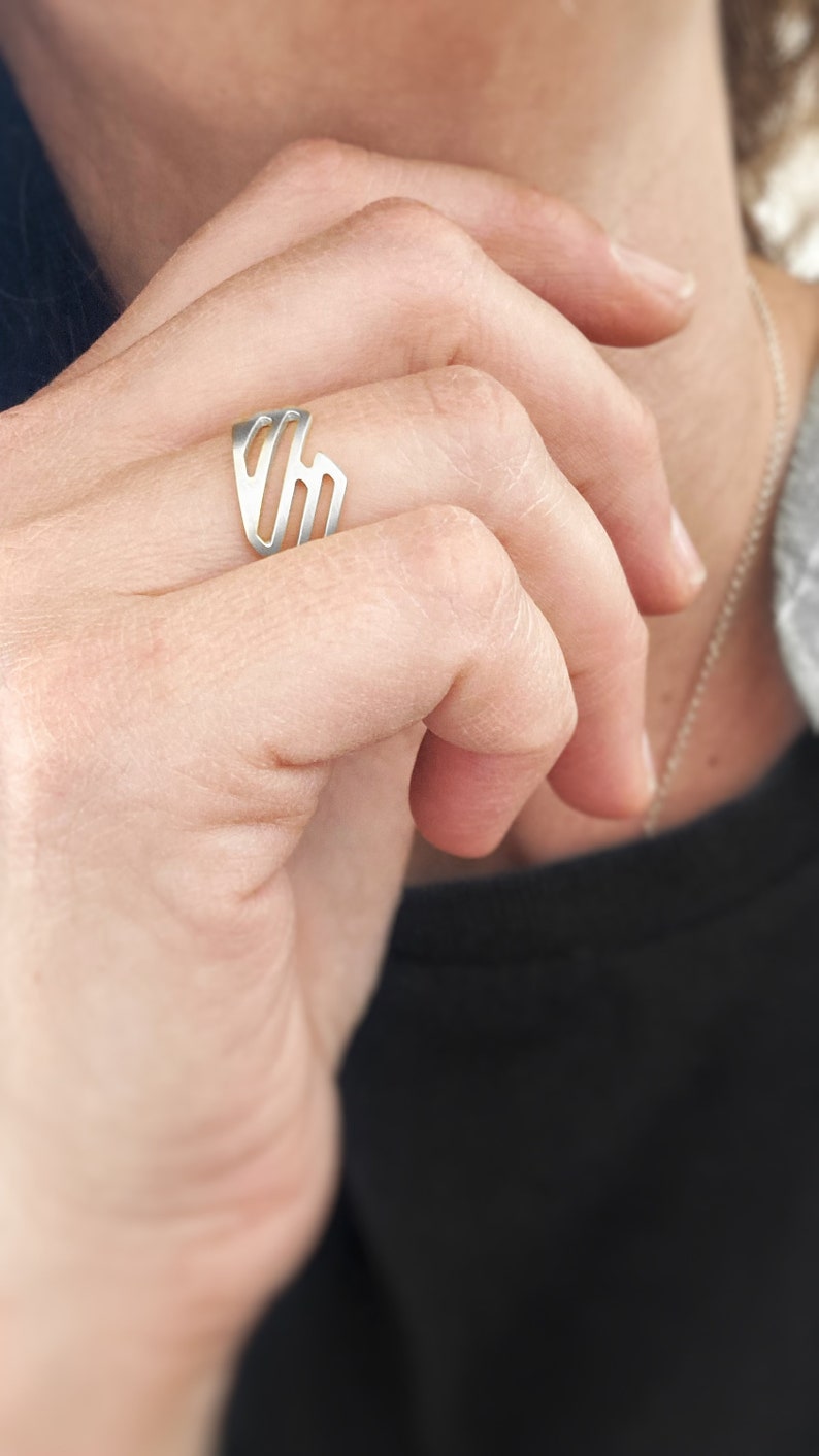 Pode incluir: Um anel de prata com um design geom&eacute;trico. O anel est&aacute; no dedo de uma pessoa.