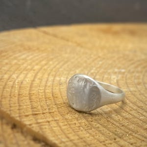 May include: A silver signet ring with a matte, textured finish on the circular face and a smooth, tapered band. The metal surface displays subtle irregularities and a brushed appearance, creating a muted, non-reflective look across the entire piece.