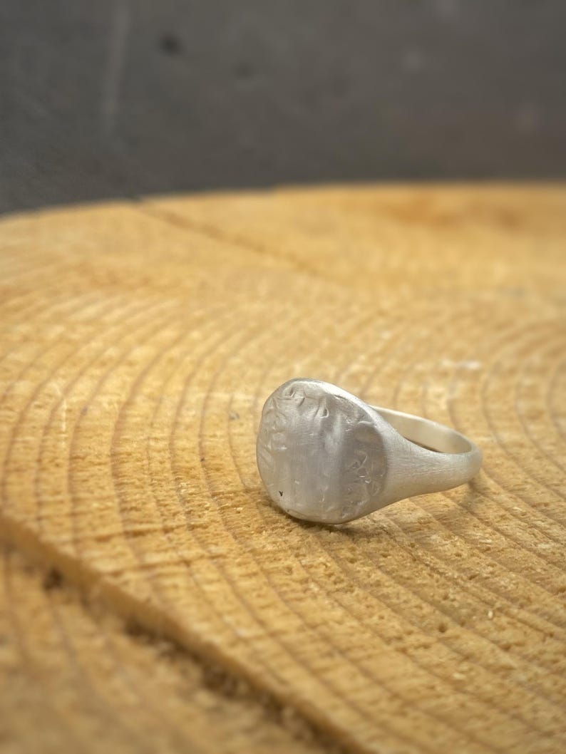 May include: A silver signet ring with a matte, textured finish on the circular face and a smooth, tapered band. The metal surface displays subtle irregularities and a brushed appearance, creating a muted, non-reflective look across the entire piece.