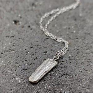 Puede incluir: Un collar plateado con un colgante rectangular texturizado. El colgante cuelga de una cadena fina. El collar se muestra sobre una superficie oscura y texturizada.