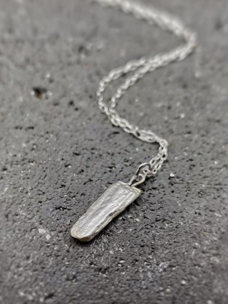 Puede incluir: Un collar plateado con un colgante rectangular texturizado. El colgante cuelga de una cadena fina. El collar se muestra sobre una superficie oscura y texturizada.