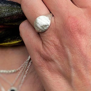 May include: A silver signet ring with a textured, uneven circular face. The metal surface has a matte, brushed finish with subtle indentations and a slightly irregular, organic shape that sits prominently on the finger of the wearer.