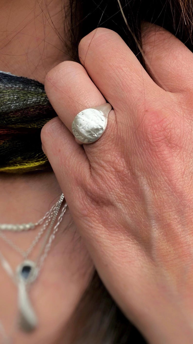 May include: A silver signet ring with a textured, uneven circular face. The metal surface has a matte, brushed finish with subtle indentations and a slightly irregular, organic shape that sits prominently on the finger of the wearer.
