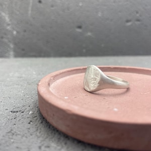 May include: A silver-toned signet ring with a textured, irregular circular face and a smooth, tapered band. The ring features a matte finish and sits on a flat, circular, terracotta-colored surface with a raised edge, showcasing its metallic luster.