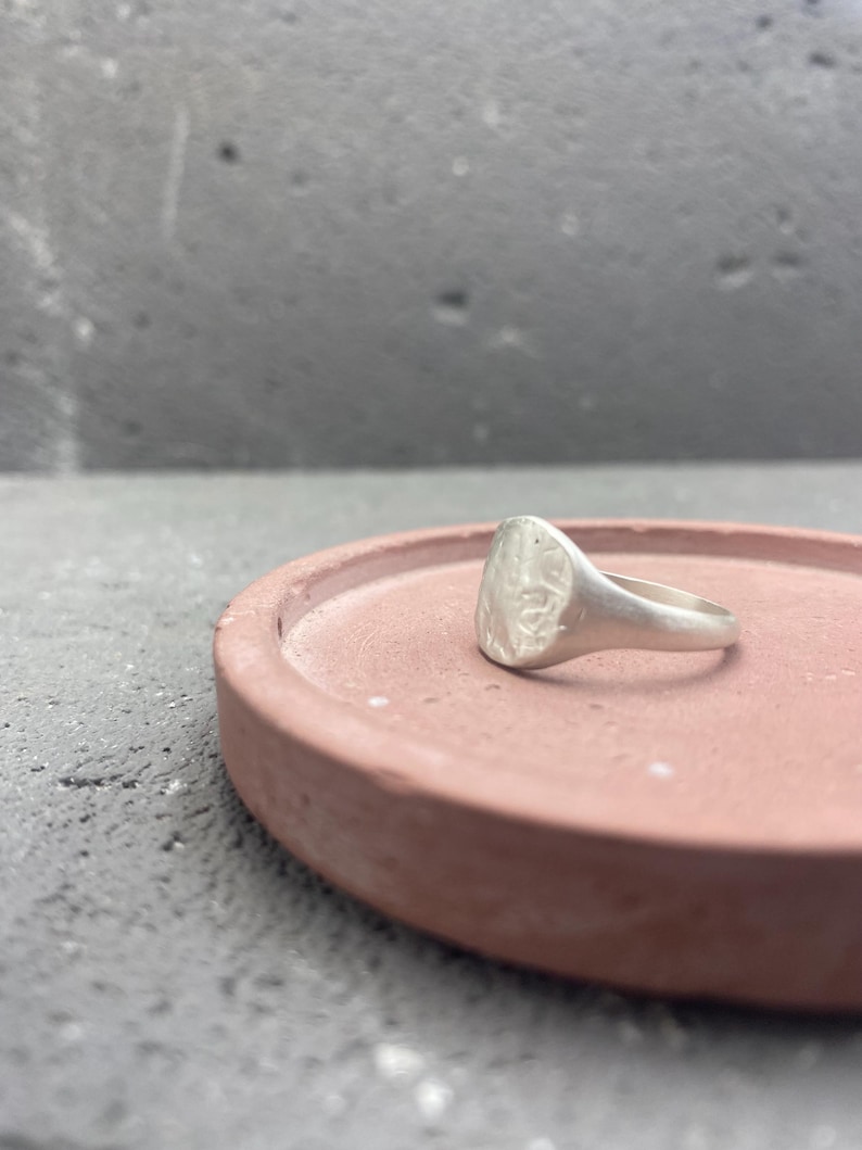 May include: A silver-toned signet ring with a textured, irregular circular face and a smooth, tapered band. The ring features a matte finish and sits on a flat, circular, terracotta-colored surface with a raised edge, showcasing its metallic luster.