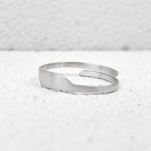 May include: A silver-colored adjustable ring with a flat, rectangular design on the front. The ring has a simple, minimalist style and is set against a white background. The ring appears to be made of metal.
