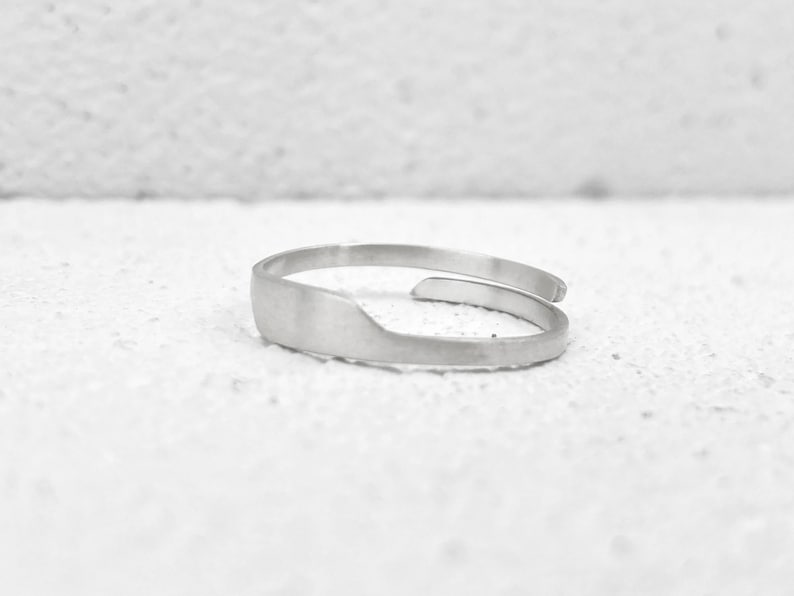 May include: A silver-colored adjustable ring with a flat, rectangular design on the front. The ring has a simple, minimalist style and is set against a white background. The ring appears to be made of metal.