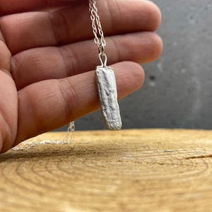 Puede incluir: Un collar de plata con un colgante rectangular texturizado. El colgante cuelga de una fina cadena. El collar se muestra sobre una superficie de madera.