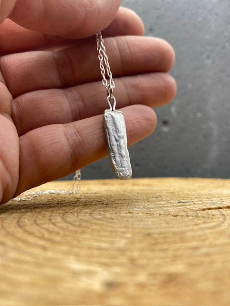 Puede incluir: Un collar de plata con un colgante rectangular texturizado. El colgante cuelga de una fina cadena. El collar se muestra sobre una superficie de madera.