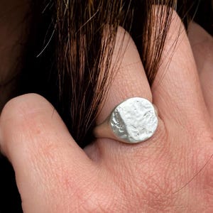 May include: A silver signet ring featuring a circular face with a textured, hammered surface. The ring has a smooth, polished band that tapers toward the back. The metal has a matte finish, highlighting the irregular, organic indentations across the top.