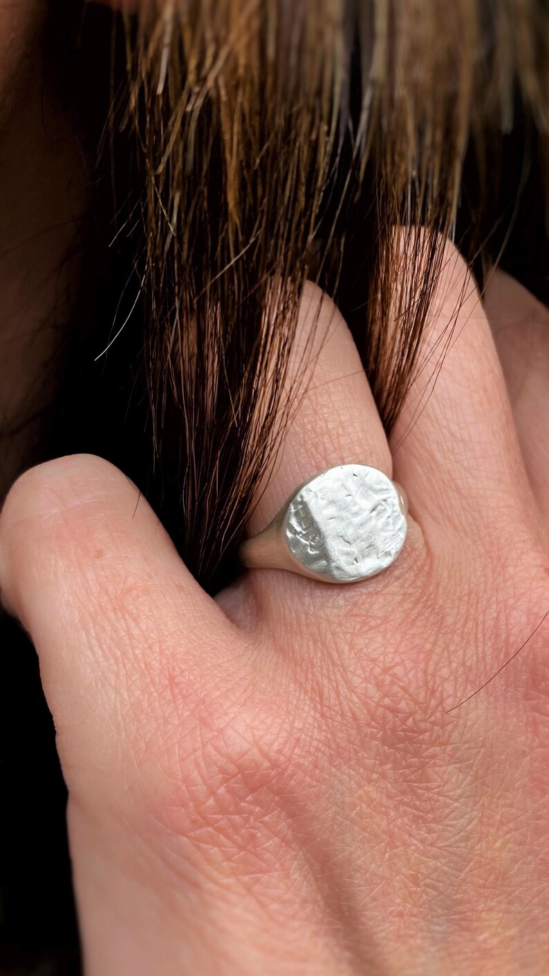 May include: A silver signet ring featuring a circular face with a textured, hammered surface. The ring has a smooth, polished band that tapers toward the back. The metal has a matte finish, highlighting the irregular, organic indentations across the top.