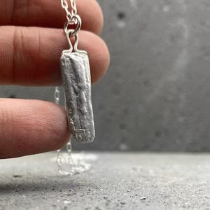 Puede incluir: Un collar de plata con un colgante rectangular con una textura que se asemeja a la corteza. El colgante cuelga de una delicada cadena de plata. El collar se muestra sobre un fondo gris.