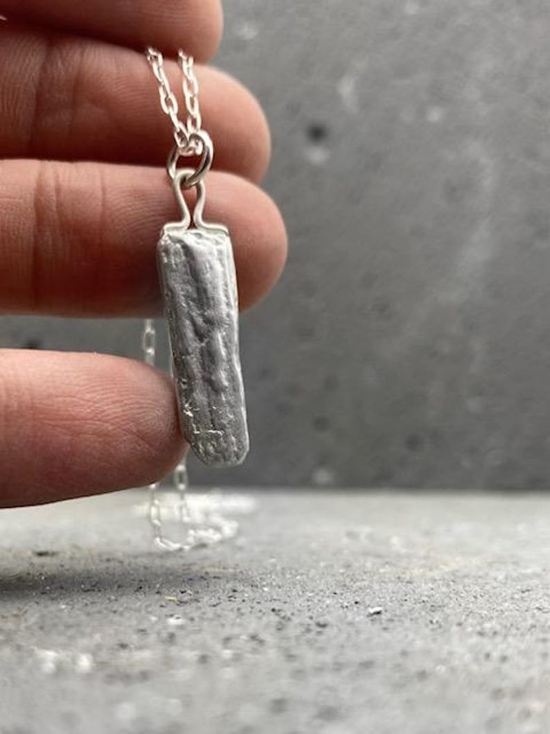 Puede incluir: Un collar de plata con un colgante rectangular con una textura que se asemeja a la corteza. El colgante cuelga de una delicada cadena de plata. El collar se muestra sobre un fondo gris.