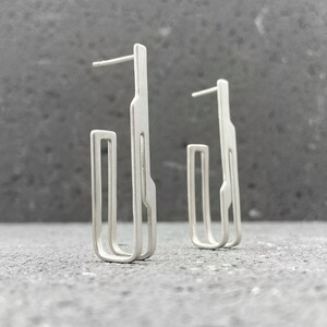May include: A pair of silver earrings with a geometric design. The earrings are made of a single piece of metal that is bent into a unique shape.