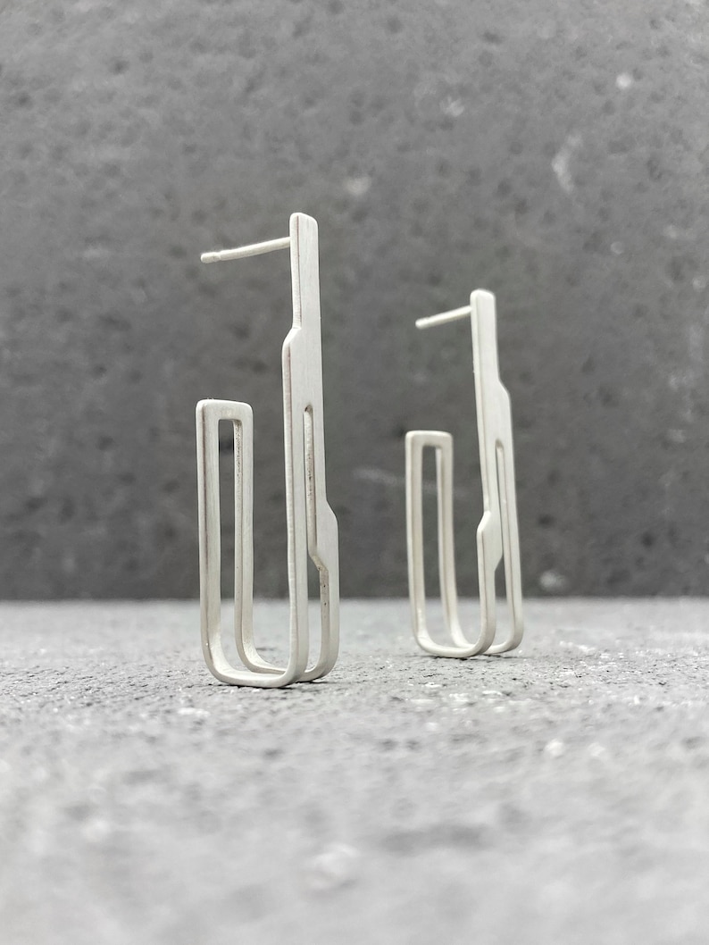 May include: A pair of silver earrings with a geometric design. The earrings are made of a single piece of metal that is bent into a unique shape.