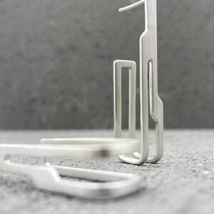 May include: A pair of silver geometric earrings with a modern design. The earrings are made of thin, flat metal and have a simple, minimalist style.