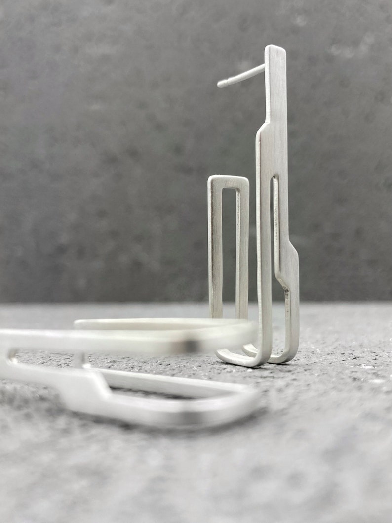 May include: A pair of silver geometric earrings with a modern design. The earrings are made of thin, flat metal and have a simple, minimalist style.
