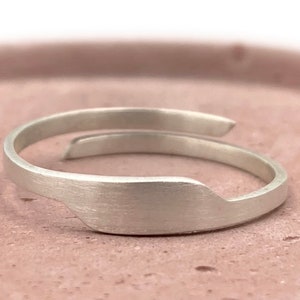 May include: A minimalist silver ring with a brushed finish. The ring features an open design with a slightly curved band that wraps around the finger. The ring's design is simple and elegant, suitable for everyday wear.
