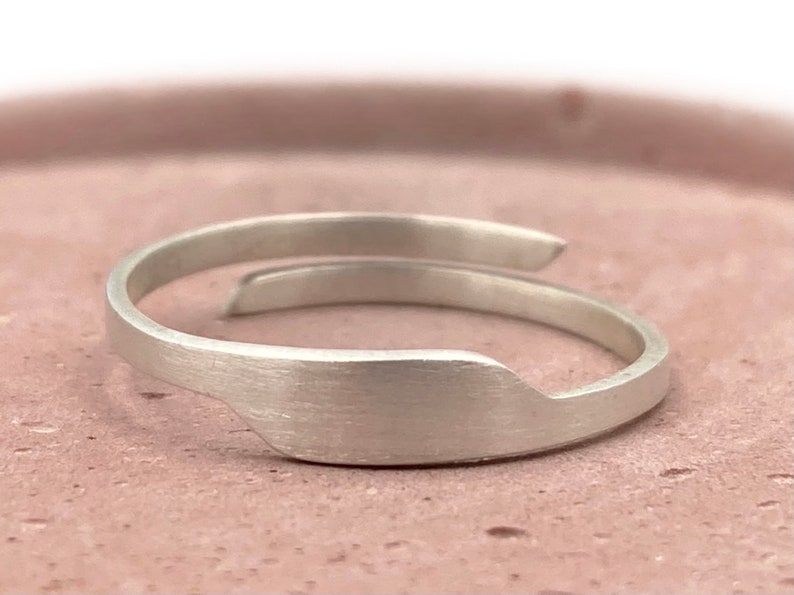 May include: A minimalist silver ring with a brushed finish. The ring features an open design with a slightly curved band that wraps around the finger. The ring's design is simple and elegant, suitable for everyday wear.