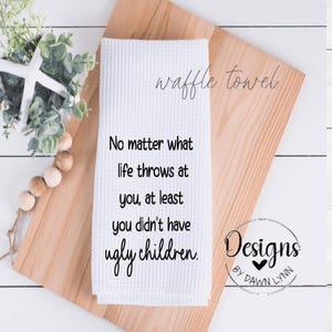 May include: White waffle towel with the text "No matter what life throws at you, at least you didn't have ugly children." The towel also has the words "waffle towel" and "Designs by Dawn Lynn."