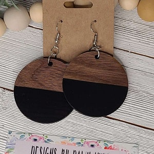May include: A pair of round wooden earrings with a black and brown wood grain pattern. The earrings are half black and half brown. The earrings are hanging from silver hooks.