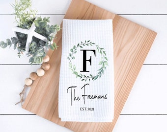 May include: A white waffle-weave kitchen towel featuring a printed design of a green watercolor leaf wreath surrounding a large black letter F. Below the wreath, the text reads The Freemans and EST. 2021 in black lettering.