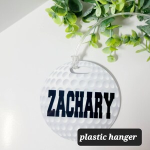 Peut inclure: &Eacute;tiquette de bagage ronde blanche avec un motif de balle de golf et le nom "ZACHARY" imprim&eacute; en noir. L'&eacute;tiquette est munie d'un crochet en plastique transparent.