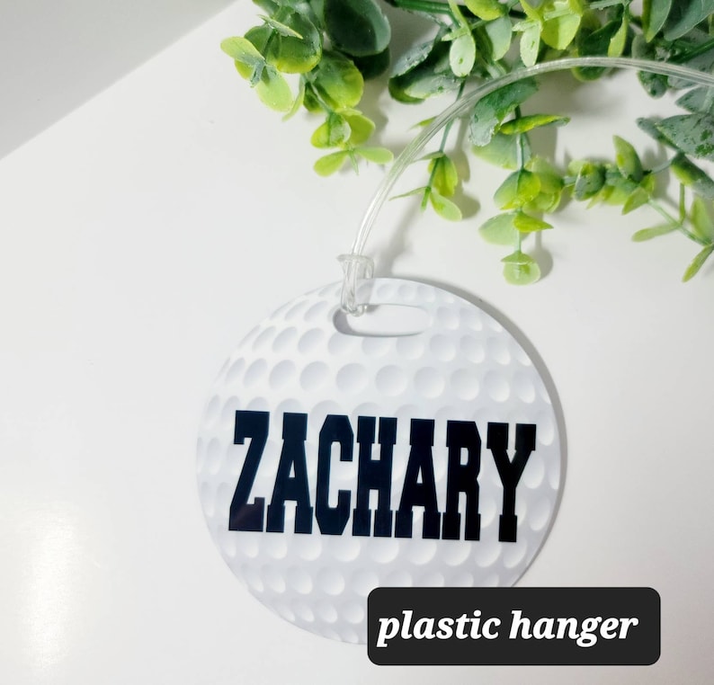 Peut inclure: &Eacute;tiquette de bagage ronde blanche avec un motif de balle de golf et le nom "ZACHARY" imprim&eacute; en noir. L'&eacute;tiquette est munie d'un crochet en plastique transparent.