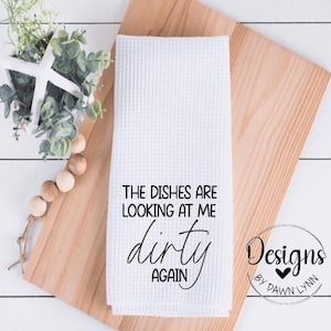 May include: White kitchen towel with black text that reads "The dishes are looking at me dirty again". The towel is on a wooden surface. The text "Designs by Dawn Lynn" is also visible in black.