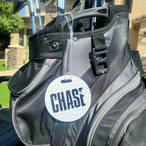 Peut inclure: Un sac de golf noir et gris avec une &eacute;tiquette ronde blanche sur laquelle est inscrit "CHASE" en lettres noires.