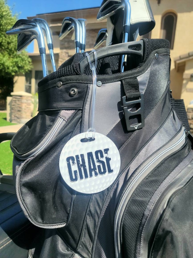 Peut inclure: Un sac de golf noir et gris avec une &eacute;tiquette ronde blanche sur laquelle est inscrit "CHASE" en lettres noires.