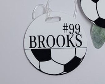 May include: White round luggage tag with a black soccer ball design. The tag has the text "#99 BROOKS" printed on it.