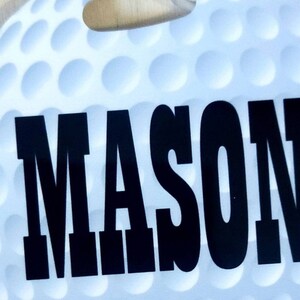 Peut inclure: Une balle de golf blanche avec une surface &agrave; picots noirs. Le nom "MASON" est imprim&eacute; en grandes lettres noires sur la balle.